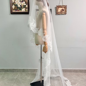 May include: A long, white bridal veil with delicate lace trim. The veil is displayed on a mannequin, showcasing its length and intricate details. The lace edges add a touch of elegance to the sheer fabric.