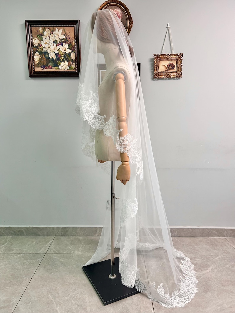 May include: A long, white bridal veil with delicate lace trim. The veil is displayed on a mannequin, showcasing its length and intricate details. The lace edges add a touch of elegance to the sheer fabric.