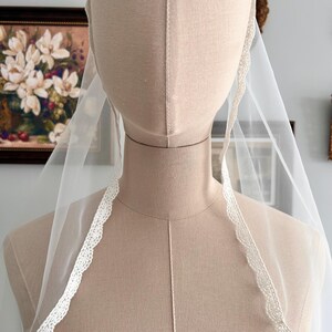 May include: A sheer white bridal veil with a delicate, scalloped lace trim. The veil is draped over a mannequin, showcasing its lightweight and flowing fabric. The lace trim adds a touch of elegance to the simple design.