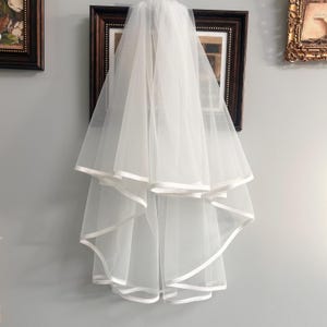 May include: A white, two-tiered bridal veil with a satin ribbon trim. The veil is made of a sheer, lightweight fabric and is designed to be worn for a wedding ceremony. The veil is hanging against a gray wall.