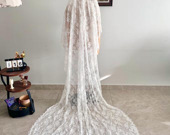 May include: A long, white lace veil with a floral pattern. The veil is draped over a person, and the train extends across the floor. The veil is a bridal accessory, and the image is taken indoors.