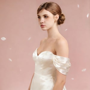 May include: A cream-colored, off-the-shoulder satin wedding dress. The bodice features a sweetheart neckline and a fitted waist. The sleeves are short and puffed. The background is a soft pink with white petal accents.