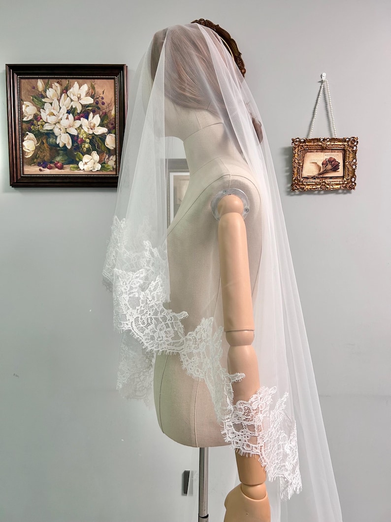 May include: A white bridal veil with delicate lace trim is draped over a mannequin. The veil is long and flowing, with intricate lace detailing along the edges. The mannequin is positioned in front of a wall with framed artwork.