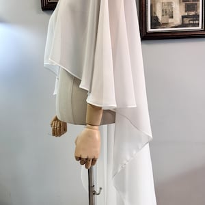 Puede incluir: Una prenda blanca y fluida estilo capa con un dobladillo alto-bajo. La tela se drapea sobre un maniqu&iacute;, mostrando la elegante silueta del dise&ntilde;o. La capa presenta mangas anchas y una espalda larga y fluida, perfecta para ocasiones formales.