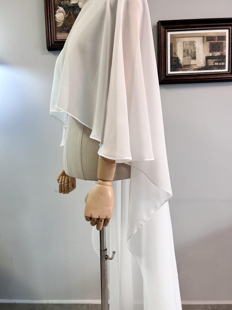 Puede incluir: Una prenda blanca y fluida estilo capa con un dobladillo alto-bajo. La tela se drapea sobre un maniqu&iacute;, mostrando la elegante silueta del dise&ntilde;o. La capa presenta mangas anchas y una espalda larga y fluida, perfecta para ocasiones formales.