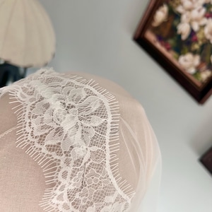 May include: Close-up of a white lace veil with intricate floral patterns and a delicate, fringed edge. The veil is made of sheer fabric, creating a soft, ethereal look. The background is blurred, focusing attention on the veil's details.