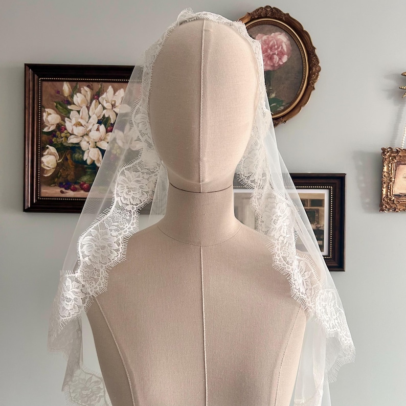 May include: A white bridal veil with delicate lace trim is displayed on a mannequin. The veil is sheer, allowing the mannequin's form to be partially visible. The lace trim adds a touch of elegance to the design. The background features framed artwork.