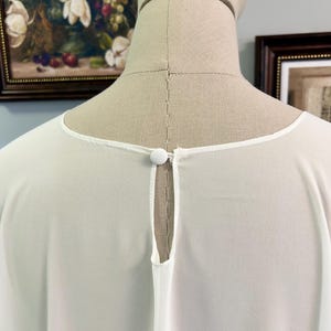 Puede incluir: Una blusa blanca y transparente con cuello redondo y detalle de ojo de cerradura en la espalda. La blusa presenta un peque&ntilde;o cierre de bot&oacute;n redondo en la parte posterior del cuello. La tela parece ligera y fluida, adecuada para climas c&aacute;lidos.