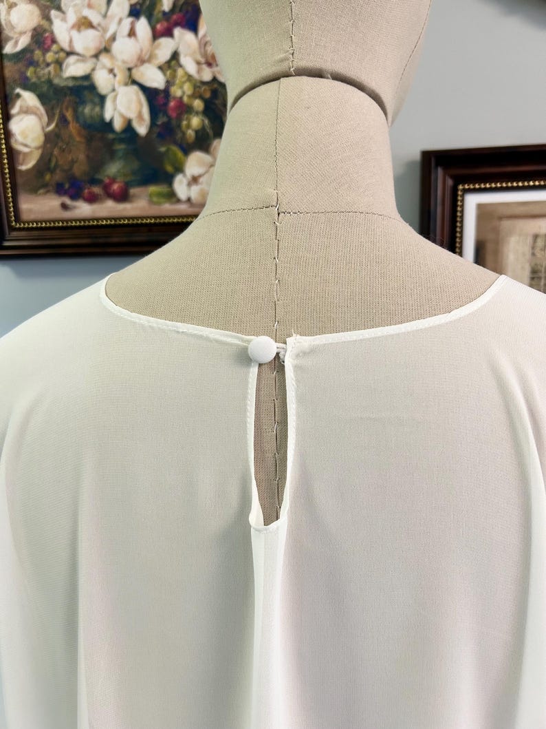 Puede incluir: Una blusa blanca y transparente con cuello redondo y detalle de ojo de cerradura en la espalda. La blusa presenta un peque&ntilde;o cierre de bot&oacute;n redondo en la parte posterior del cuello. La tela parece ligera y fluida, adecuada para climas c&aacute;lidos.