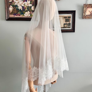 Pode incluir: Um véu de noiva branco transparente com uma delicada barra de renda. O véu é drapeado sobre um manequim, mostrando seu comprimento e design intrincado. A barra de renda adiciona um toque de elegância ao acessório de noiva.