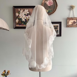 May include: A white bridal veil with a delicate lace trim, displayed on a mannequin. The veil is sheer, allowing light to pass through, and features a scalloped lace edge. The veil is a classic accessory for weddings.