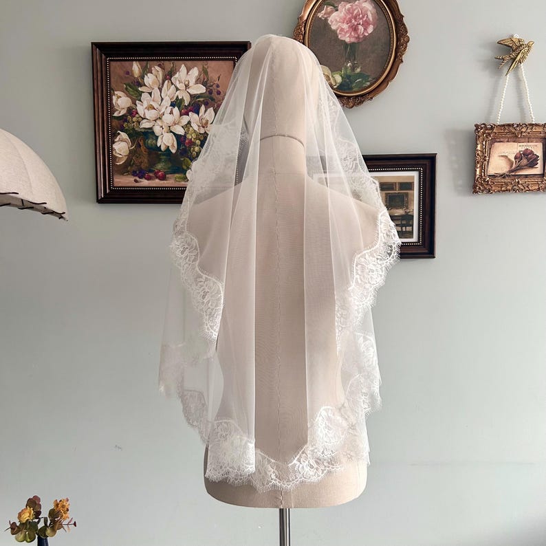 May include: A white bridal veil with a delicate lace trim, displayed on a mannequin. The veil is sheer, allowing light to pass through, and features a scalloped lace edge. The veil is a classic accessory for weddings.
