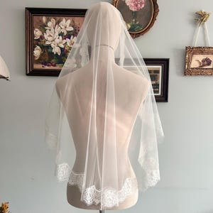 May include: A sheer, white bridal veil with delicate lace trim. The veil is draped over a mannequin, showcasing its soft texture and design. The lace detailing adds a touch of elegance to the wedding accessory.