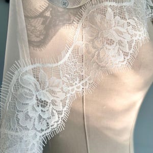 May include: Close-up of white floral lace trim with a scalloped edge. The delicate lace features a detailed floral pattern and is set against a soft, neutral background. The trim is likely used for embellishing garments or crafts.