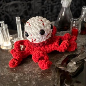 Peut inclure: Un jouet poulpe fait à la main au crochet, avec une tête blanche ornée de touches de rouge et de grands yeux noirs. Le poulpe a un corps et des tentacules rouges. Le jouet est présenté sur une surface texturée avec des bouteilles en verre en arrière-plan.