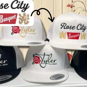 May include: White and black baseball caps with embroidered text and graphics. The white caps feature "Rose City Banquet" and "Tyler TEXAS" with a rose. The black caps have similar designs. The caps have a flat brim.