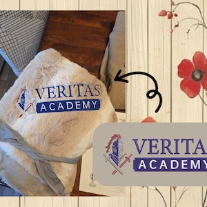 May include: A plush, cream-colored blanket with the embroidered text "VERITAS ACADEMY" in navy blue. The blanket is folded and tied with a gray ribbon. The image also shows a logo with the same text and a knight's helmet and crossed swords.