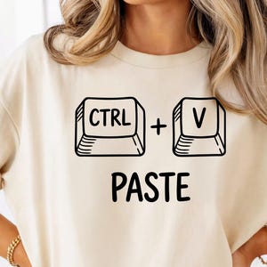May include: A beige t-shirt featuring a black graphic of the CTRL and V keys with a plus sign in between, and the word "PASTE" underneath. The shirt has short sleeves and a crew neck.