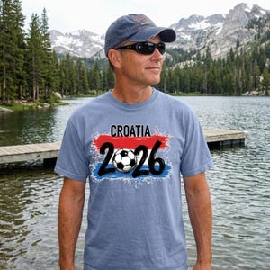 May include: A light blue t-shirt with the word "CROATIA" above the number "2026" and a soccer ball graphic. The design features red and blue accents, and the shirt is worn by a person wearing sunglasses and a baseball cap.