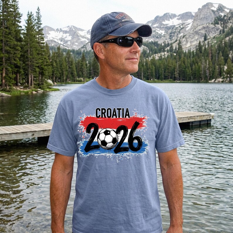 May include: A light blue t-shirt with the word "CROATIA" above the number "2026" and a soccer ball graphic. The design features red and blue accents, and the shirt is worn by a person wearing sunglasses and a baseball cap.