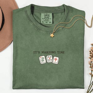 a green t-shirt with the text "IT'S MAJJONG TIME" printed on it, along with a pair of dominoes and a star pendant. The t-shirt is laid out on a white surface, and there is a brown hat and some flowers nearby.