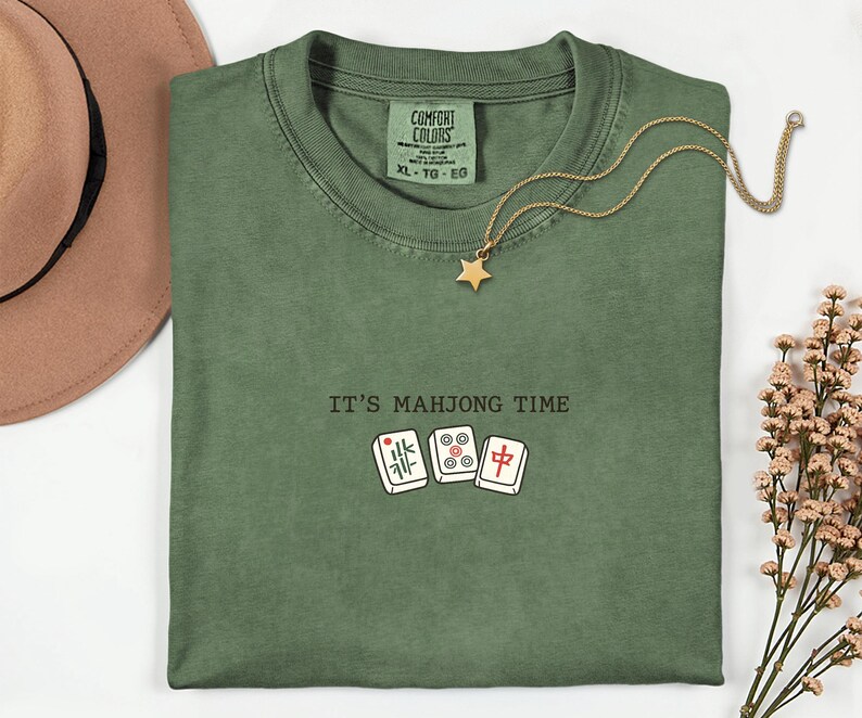 a green t-shirt with the text "IT'S MAJJONG TIME" printed on it, along with a pair of dominoes and a star pendant. The t-shirt is laid out on a white surface, and there is a brown hat and some flowers nearby.