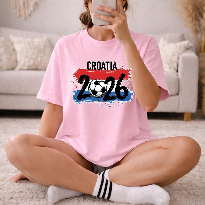 May include: A light pink t-shirt with the word "CROATIA" in black letters above the numbers "2026" and a soccer ball. The numbers and soccer ball are in black. The design has red and blue brush strokes.