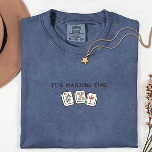 a blue t-shirt with the text "IT'S MAHJONG TIME" printed on it, along with a pair of Chinese tiles. The t-shirt is laid out on a white surface, and there is a hat and some flowers nearby.
