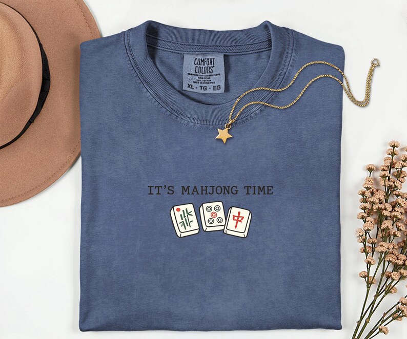 a blue t-shirt with the text "IT'S MAHJONG TIME" printed on it, along with a pair of Chinese tiles. The t-shirt is laid out on a white surface, and there is a hat and some flowers nearby.