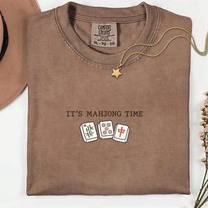 a brown t-shirt with the text "IT'S MAJONG TIME" printed on it, along with a pair of dominoes and a star pendant.
