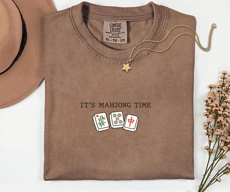 a brown t-shirt with the text "IT'S MAJONG TIME" printed on it, along with a pair of dominoes and a star pendant.