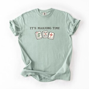 A light green t-shirt with the text "IT'S MAHJONG TIME" printed on it, along with illustrations of playing cards.