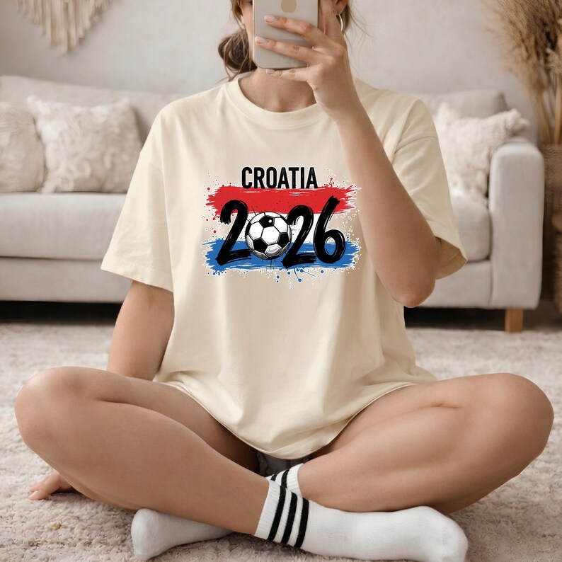 May include: Cream-colored t-shirt with the word "CROATIA" above the numbers "2026" and a soccer ball graphic. The design incorporates red, white, and blue brushstrokes, resembling a flag. The shirt is a casual, short-sleeved style.