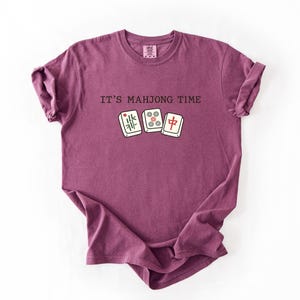 A purple t-shirt with the text "IT'S MAJONG TIME" printed on it, along with a graphic of a mahjong tile.