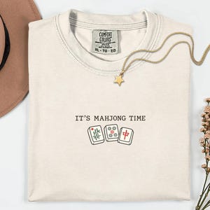 a white t-shirt with the text "IT'S MAJJONG TIME" printed on it, along with a pair of dominoes and a star charm. The t-shirt is laid out on a white surface, with a brown hat and some flowers nearby.