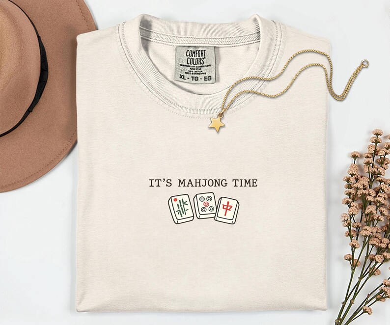 a white t-shirt with the text "IT'S MAJJONG TIME" printed on it, along with a pair of dominoes and a star charm. The t-shirt is laid out on a white surface, with a brown hat and some flowers nearby.