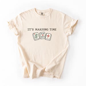 A beige t-shirt with the text "IT'S MAJONG TIME" printed on it, along with illustrations of dominoes.