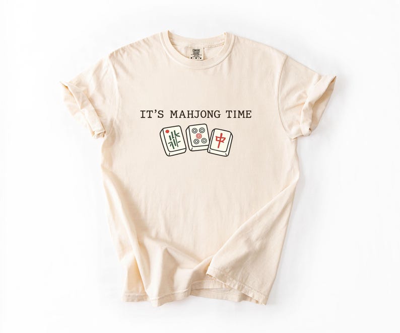 A beige t-shirt with the text "IT'S MAJONG TIME" printed on it, along with illustrations of dominoes.