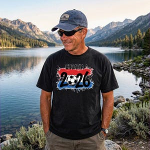 May include: Black t-shirt with the word "CROATIA" above the number "2026" and a soccer ball graphic. The text is in white, red, and blue. The person is wearing a blue cap and sunglasses.