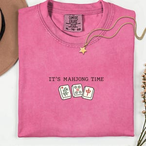 a pink t-shirt with the text "IT'S MAJONG TIME" printed on it, along with a pair of dominoes and a star pendant.