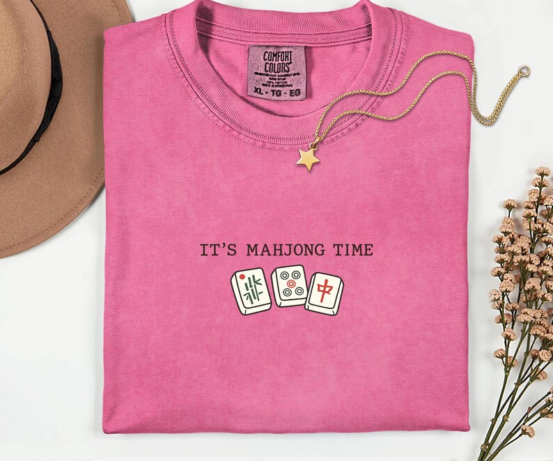 a pink t-shirt with the text "IT'S MAJONG TIME" printed on it, along with a pair of dominoes and a star pendant.