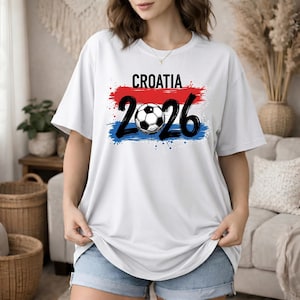May include: White t-shirt with the word "CROATIA" in black letters above the numbers "2026" with a soccer ball in the center. The text is set against a red and blue paint stroke design.