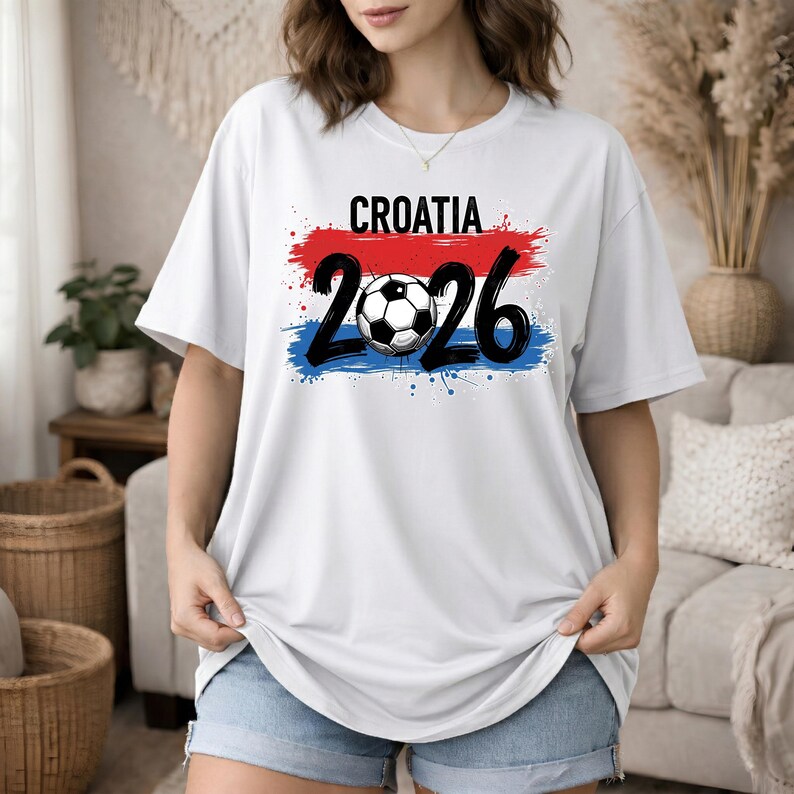 May include: White t-shirt with the word "CROATIA" in black letters above the numbers "2026" with a soccer ball in the center. The text is set against a red and blue paint stroke design.