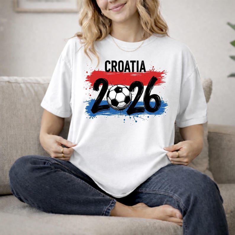 May include: White t-shirt with the word "CROATIA" above the numbers "2026" and a soccer ball graphic. The design incorporates red, white, and blue brushstrokes, resembling a flag. The shirt is made of a soft material.