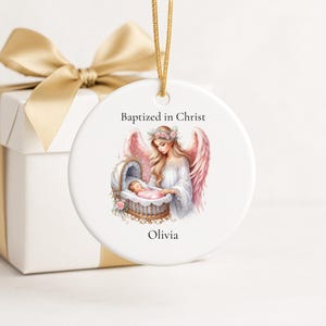 May include: A round white ceramic ornament hanging by a gold cord. The center features an illustration of an angel with pink wings watching over a baby in a cradle. Text reads Baptized in Christ and the name Olivia in black lettering.