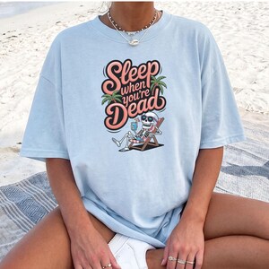 May include: A light blue t-shirt featuring a graphic of a skeleton lounging in a beach chair under a palm tree. The skeleton wears heart-shaped sunglasses and holds a drink. The text reads, Sleep when you're DEAD in a stylized pink font.