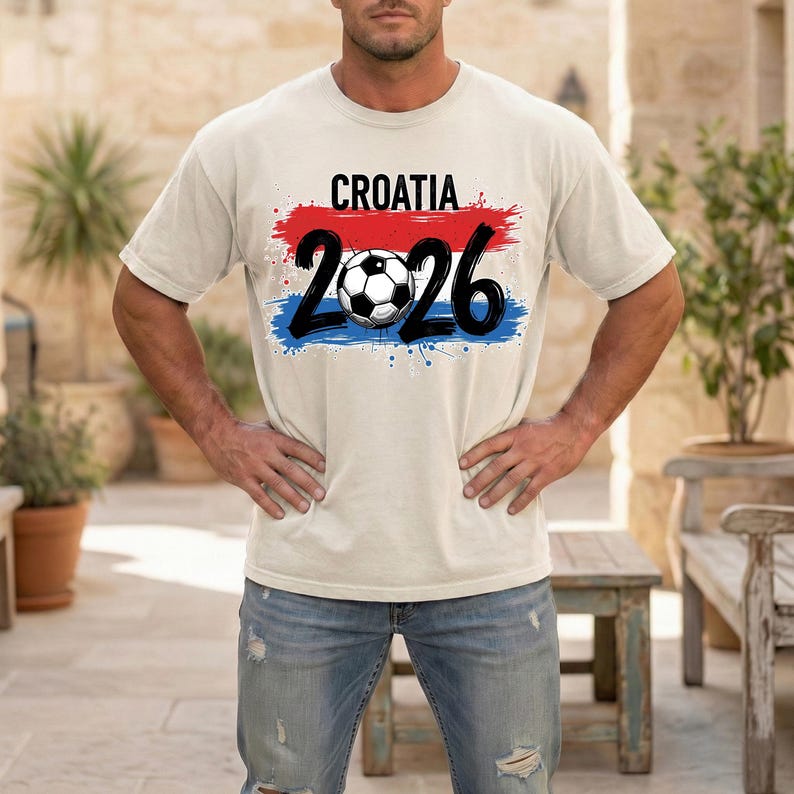 May include: A light beige t-shirt with the word "CROATIA" in black above the number "2026" and a soccer ball graphic. The design features red and blue paint strokes. The shirt is made of a soft material.