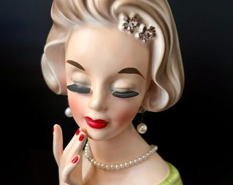 Peut inclure: Un cache-pot en céramique représentant une figure stylisée aux cheveux blonds, vêtue d'un vêtement vert et portant un collier de perles. La figure a les yeux fermés avec de longs cils noirs, des lèvres rouges et des ongles rouges.