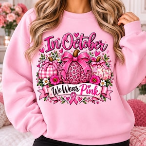 May include: A light pink sweatshirt featuring a graphic print of three pumpkins decorated with pink bows, leopard print, and plaid patterns. The design includes floral accents, small hearts, and the text In October We Wear Pink with a breast cancer ribbon.