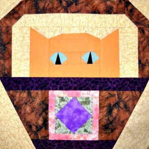 May include: A geometric quilt block featuring an orange cat face with blue eyes and black pupils. The cat is set against a tan background, with a purple and brown patterned border. A square with a purple diamond is at the bottom.